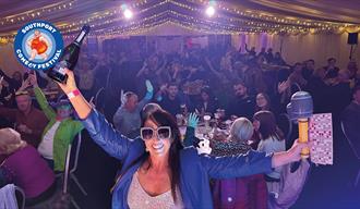 Image of a lady smiling wearing sunglasses and a sparkly top at Comedy Bingo holding a bottle of bubbly in one hand and a bingo card and blow up micro