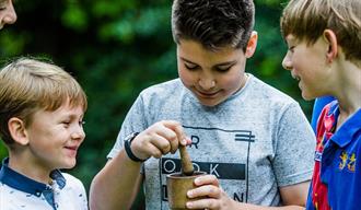 child mixes materials in a wooden pot surrounded by other children outdoors