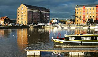 A colour photograph of Gloucester Docks including warehouse buildings, the water basin and a narrowboat in the foreground.