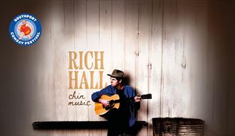 Image of American Comedian Rich Hall sitting down playing the guitar and looking to his right, his name and the name of his show Chin music are writte