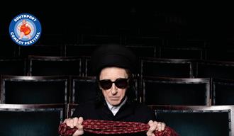 image of Dr John Cooper Clarke wearing dark sunglasses and a black hat sitting in an empty theatre looking to camera holding a red scarf