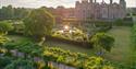 Aerial view of a historic mansion surrounded by manicured gardens. A fountain glows in sunlight, with lush trees and hedges under a serene sky.