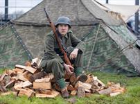 A soldier in WW2 uniform holding a rifle and sitting on a pile of split logs at the Weston-super-Mare Helicopter Museum Military History weekend