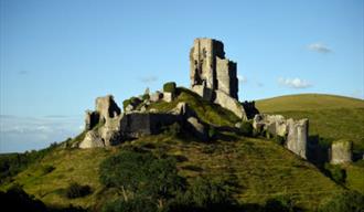 The ruins of Corfe Castle stand high a top a grass covered hill dominating the landscape below. The sky is blue and the sun is shining