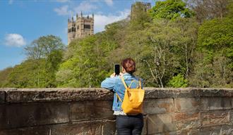 A person takes a photo of Durham Cathedral from a bridge among the wooded riverbanks