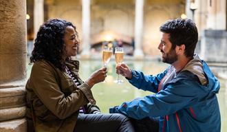 A man and woman toasting with champagne glasses in front of the Great Bath at the Roman Baths