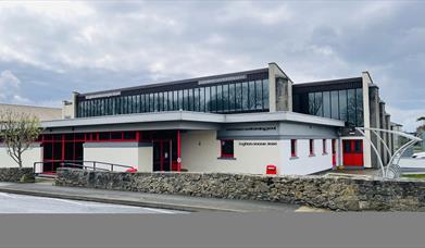 Cream coloured swimming pool building with red doors and windows. Glazing over the first floor.