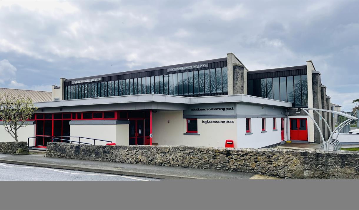 Cream coloured swimming pool building with red doors and windows. Glazing over the first floor.