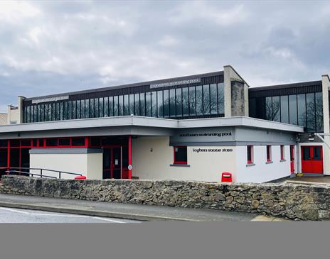 Cream coloured swimming pool building with red doors and windows. Glazing over the first floor.