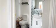 Modern bathroom with a white toilet, wicker shelving unit stocked with toilet rolls, and a white vanity unit with a backlit mirror. The wall behind th