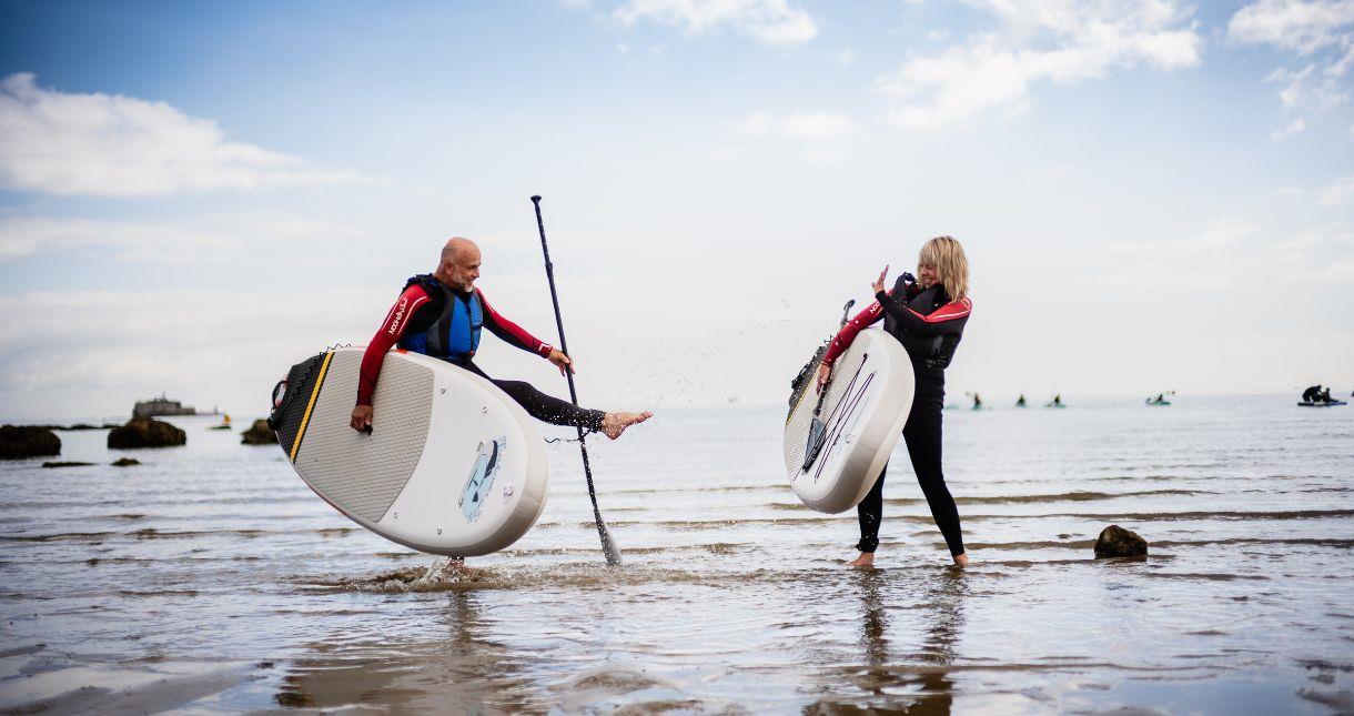 Couple holding paddle boards and spraying sea water at each other