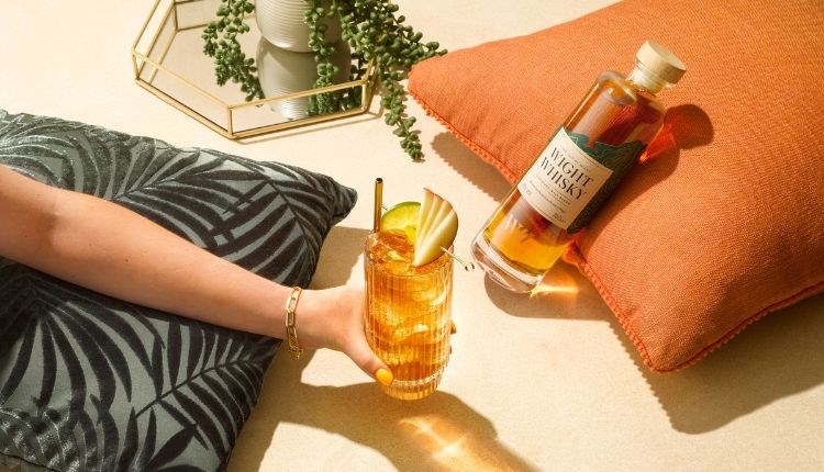 Lady holding a drink and a Wight Whisky bottle is laying by the side of the drink on a cushion, local produce, Isle of Wight