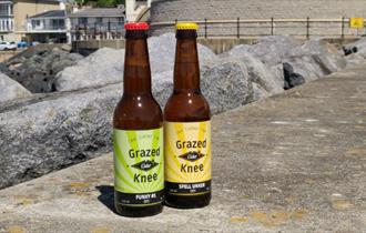 Two bottles of Grazed Knee Cider bottles standing on the walkway at Ventnor Esplanade, Isle of Wight, local producers, local produce, let's buy local