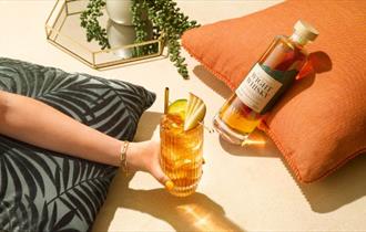 Lady holding a drink and a Wight Whisky bottle is laying by the side of the drink on a cushion, local produce, Isle of Wight