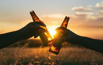 Two people clunking two bottles of beer together with a sunset behind, Beer, music and poetry festival at Waverley Park Holiday Centre, Isle of Wight,