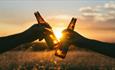 Two people clunking two bottles of beer together with a sunset behind, Beer, music and poetry festival at Waverley Park Holiday Centre, Isle of Wight,