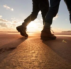 Couple wearing walking boots walking along beach with sunset in background