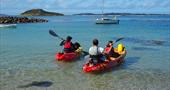 A family leaving the beach in a single and double kayak. They are kayaking on calm, crystal clear water.
