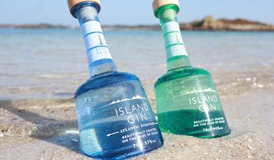 Picture of an aqua bottle of Island Gin and an azure blue bottle Atlantic Strength in the sand.