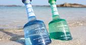 Picture of an aqua bottle of Island Gin and an azure blue bottle Atlantic Strength in the sand.