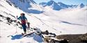 Picture of a ski hiker skiing up a mountain side. Mountains covered in snow in the backdrop.