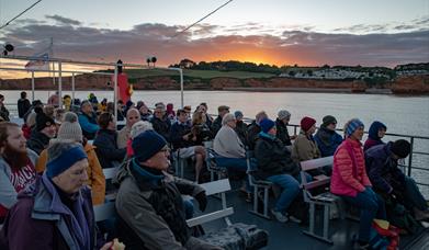Picture featuring a sunset in the back ground and the top level of a boat trip with people looking out at the scenery