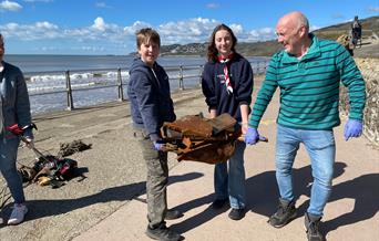 Picture taken beside the beach with 3 people carrying some rusty rubbish wearing gloves