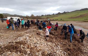 Photo showing lots of people on a beach next to a river looking for rubbish - cloudy skies, some dogs on the leads