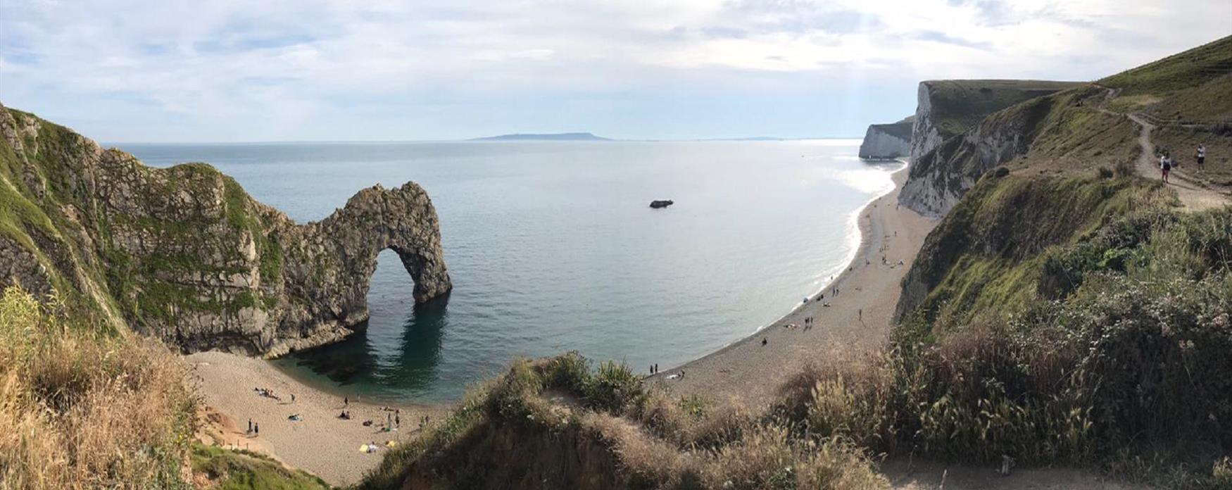 A wide angled view of the coastal arch of Durdle Door, the beach and the rolling coastal cliffs beyond.