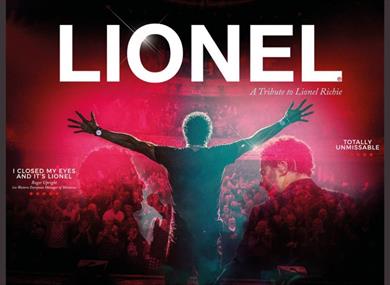 The performer is standing on a stage with his back to the camera looking out to the audience with arms stretched.