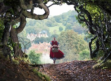 A lady in a red dress carries a basket through the woods with a the sunny countryside behind her on the horizon.