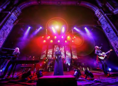 The performers are on stage with a singer in the centre in a long black gown.  The stage is bathed in blue, purple and orange lights.