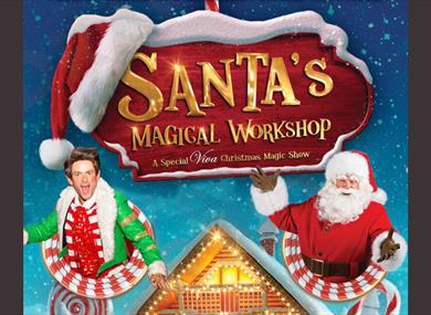 Santa and an elf sit under a snow covered sign for Santa's Magical Workshop.