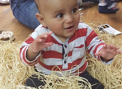 A happy toddler is sat amongst straw like material, reaching out to touch it.