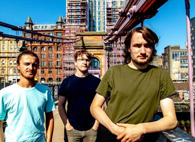 A photograph of the three performers, standing on a bridge with a city landscape in the background.