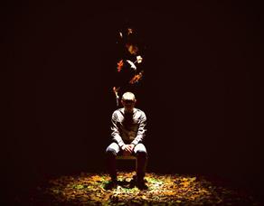 Image credit Drew Forsyth.  A man sit on a chair on the stage surrounded in darkness with the spotlight on him.  Autumn leaves fall from above and are