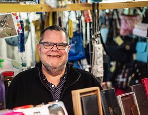 A male stallholder wearing glasses smiles at the camera from behind the table of wallets and purses for sale