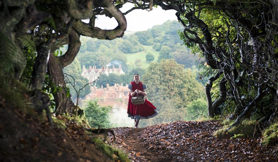 A lady in a red dress carries a basket through the woods with a the sunny countryside behind her on the horizon.