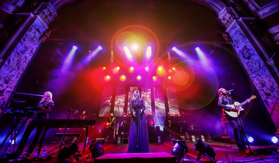 The performers are on stage with a singer in the centre in a long black gown.  The stage is bathed in blue, purple and orange lights.