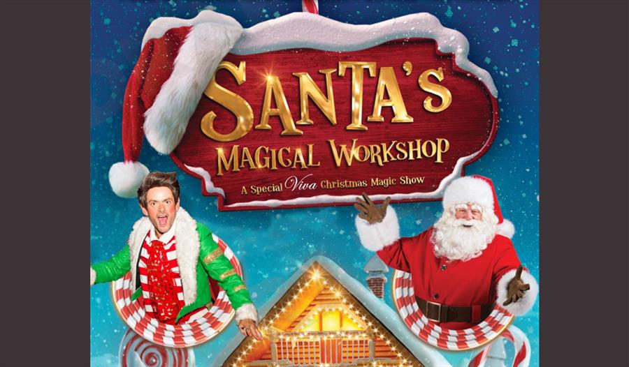 Santa and an elf sit under a snow covered sign for Santa's Magical Workshop.
