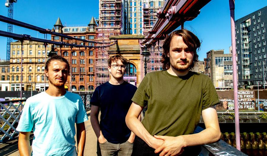 A photograph of the three performers, standing on a bridge with a city landscape in the background.