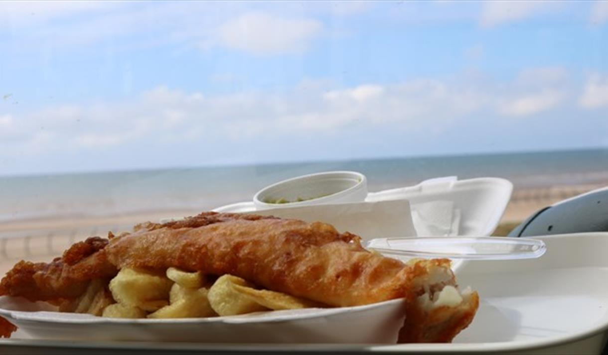 A plate of fish and chips against the backdrop of Blackpool beach on a sunny day.