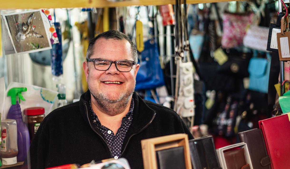A male stallholder wearing glasses smiles at the camera from behind the table of wallets and purses for sale