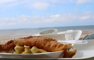 A plate of fish and chips against the backdrop of Blackpool beach on a sunny day.