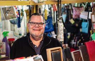 A male stallholder wearing glasses smiles at the camera from behind the table of wallets and purses for sale