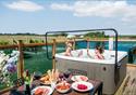 An inviting spread of food sits on a patio dining table.  Behind two ladies enjoy a hot tub, with a glass of fizz.