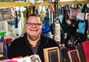 A male stallholder wearing glasses smiles at the camera from behind the table of wallets and purses for sale