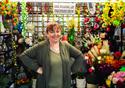 A woman with her hands on her hips smiles broadly at the camera. Surrounding her are a range of artificial flowers she has for sale