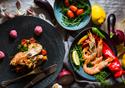 A selection of seafood dishes sit on a table surrounded by lemon, peppers, onions and garlic ingredients.