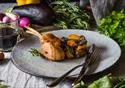 A chicken dish sits on a table surrounded by herbs, vegetables, a wine cork and a glass of red.